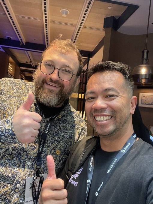 Two smiling men pose together for a selfie, both giving thumbs up; one wears a patterned blue paisley shirt with glasses and a 'Tokyo Web3' lanyard, while the other wears a gray shirt and 'Bitmarkets' lanyard.