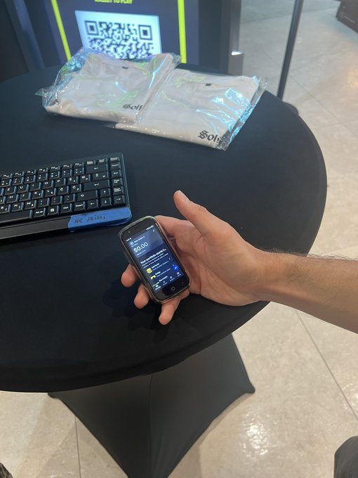 A hand holds a portable payment device displaying a $0.00 prompt on a black table, with a keyboard, packaged items, and a QR code stand nearby.