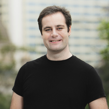 A smiling man with short brown hair wears a plain black t-shirt, standing outdoors with a blurred building in the background.