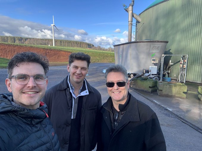 Outdoor selfie photo of three men standing in front of an industrial biogas facility, with a wind turbine and green hills visible in the background under a partly cloudy sky.