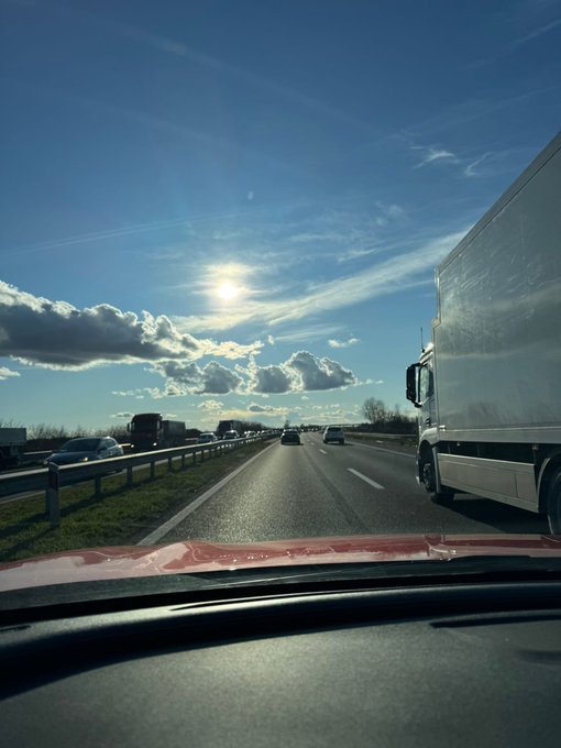 A view from inside a car driving on a highway under a bright sun and blue sky with scattered clouds, with a white truck visible on the right and other vehicles ahead.