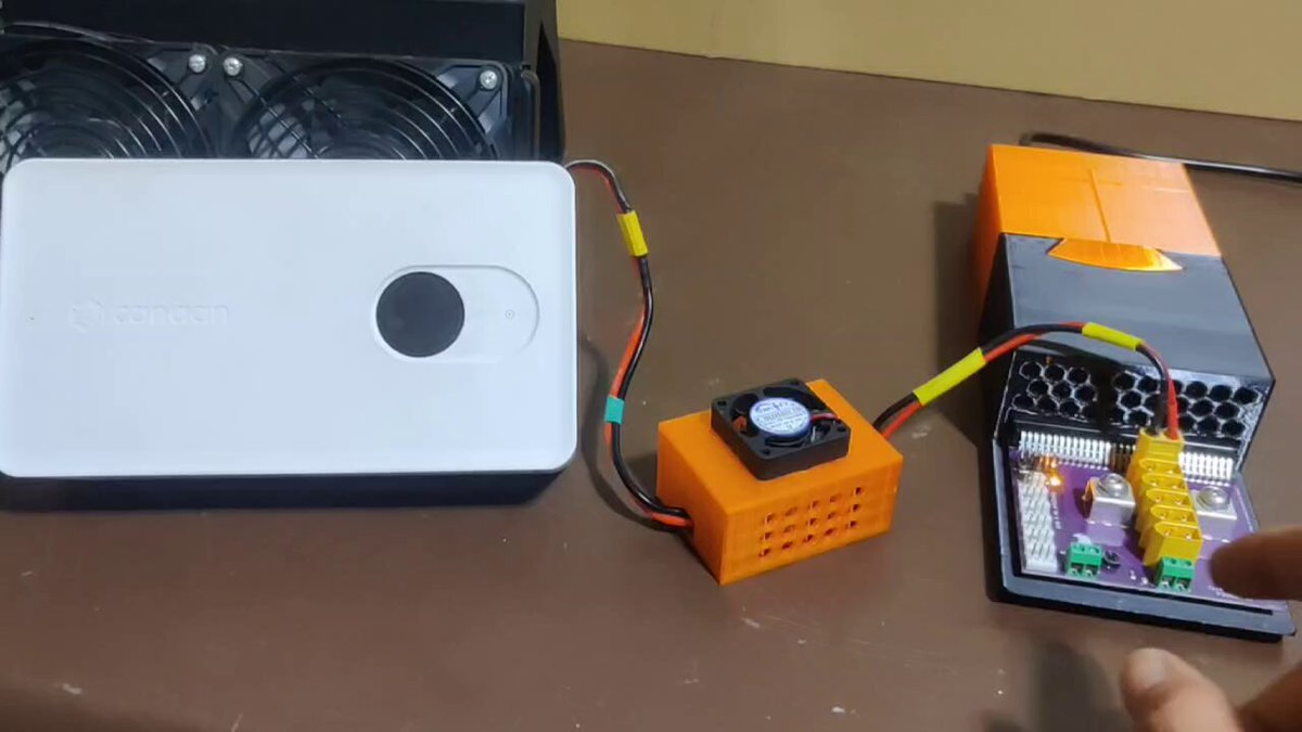 The image shows a person's hand holding a small electronic device, possibly a circuit board or a component of some sort. The device appears to be connected to an electronic device that has a white box with a black screen attached to it. The background is blurred, but there are two other devices in the scene - one on the left side and another on the right side.