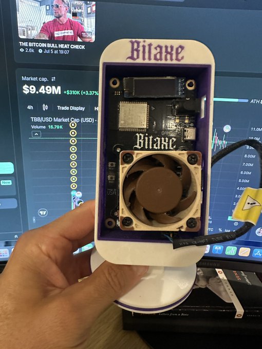 A hand holds a BitAxe open-source Bitcoin solo mining device, with the computer screen behind displaying a market cap of $9.49M and a YouTube video titled "THE BITCOIN BULL HEAT CHECK" showing 2.6K views, illustrating the post's request to give away BitAxe devices to Bitcoin Bull community members for home solo mining.