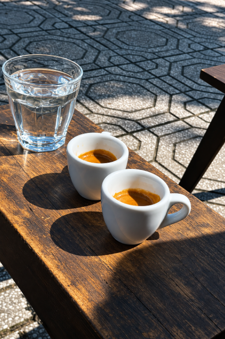 Two small white espresso cups filled with coffee and a glass of water sit on a wooden table outdoors, casting strong shadows on the stone-patterned pavement below.