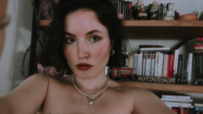 A softly lit portrait of a person with short dark hair and dark lipstick wearing a pearl necklace, with a bookshelf filled with books and small objects visible in the background.