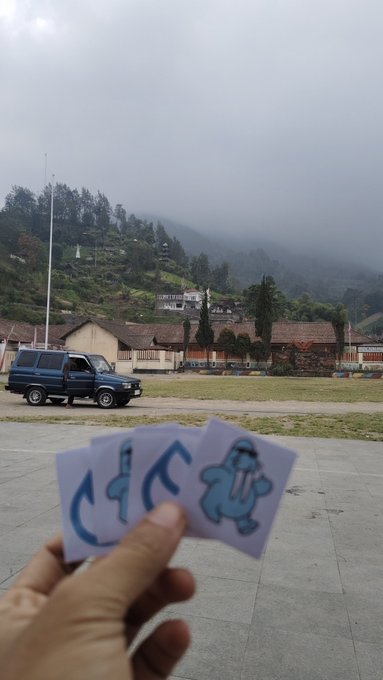 A hand holding three small cards with blue graffiti-style lettering ('CCC' and an alien) against a backdrop of a foggy, mountainous village with a parked SUV.