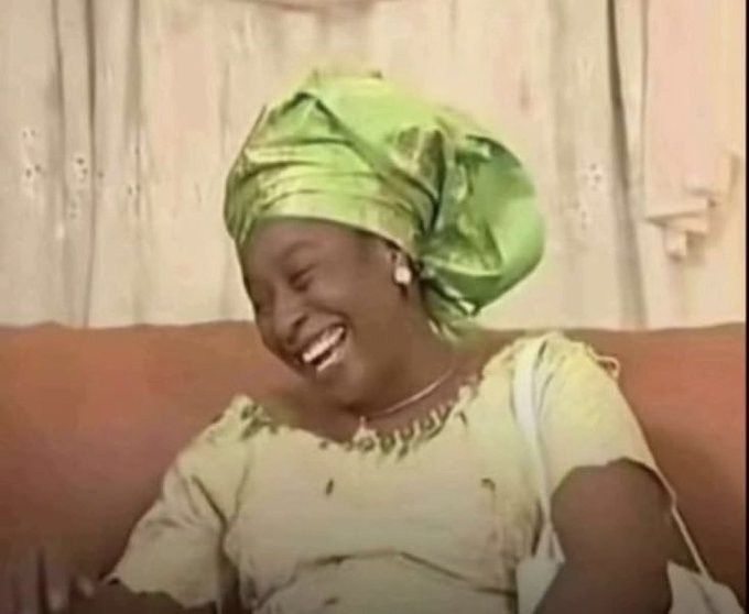 A woman wearing a shiny green headwrap and matching yellow-green traditional outfit laughs heartily while seated on a brown couch.