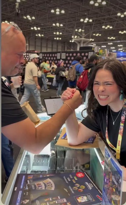 A candid photo at what appears to be a convention, showing a woman with a strained smile arm-wrestling a man at a vendor booth, with crowds and tables visible in the background.