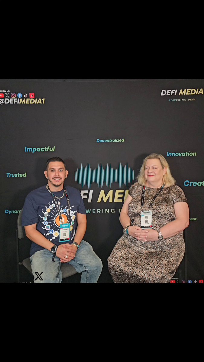 In this image, a man and woman are sitting next to each other on chairs, both wearing name tags that indicate they are participating in an interview or panel discussion. The backdrop of the scene features a black wall with white text, which includes words such as "Define Media", "FIRE MINE", "INNOVATION", "TRUSTED", and "CREATIVITY". The woman is wearing a brown dress while the man has on a blue shirt. They are both smiling for the camera, suggesting they are enjoying the event or conversation.
