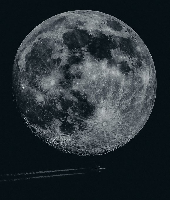 A detailed grayscale image shows a full moon with visible craters and surface texture against a dark background, with a small aircraft contrail visible at the bottom.