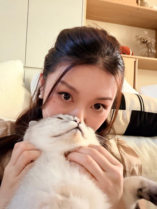 A young woman with her hair pulled back cuddles a white cat that's tilted upward with its eyes closed in apparent bliss, set in a cozy room with shelving and a small flower arrangement in the background.