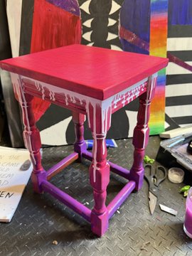 A small wooden side table with its top half painted red and bottom half purple, with white paint dripping between the two colors, sitting in what appears to be an art studio.