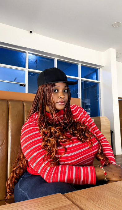 A young woman with long auburn braids, wearing a black cap and red-and-white striped shirt, sitting at a booth in what appears to be a restaurant or cafe.