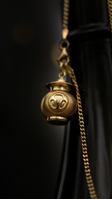 A bronze-colored spherical pendant featuring a Triforce symbol hangs from a golden chain against a dark fabric background.