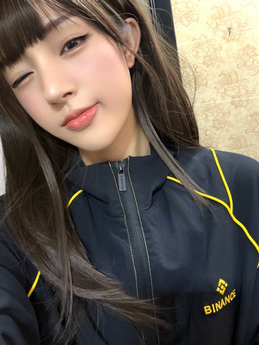A selfie of a woman with long dark hair and bangs winking at the camera while wearing a black and yellow Binance-branded jacket.