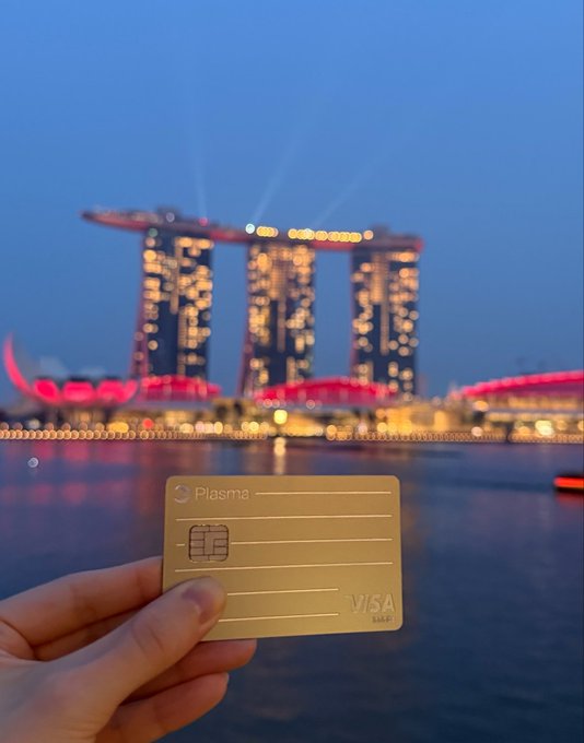 A hand holds a gold 'Plasma VISA' card in front of the illuminated Marina Bay Sands hotel in Singapore at twilight.