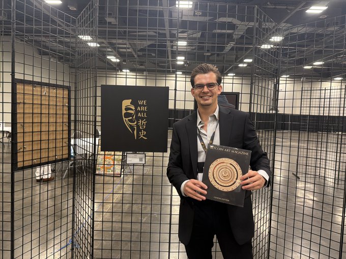 A smiling man in a black suit holds a 'Wiccan Art Magazine' in front of a wire mesh display at what appears to be a convention booth with a 'WE ARE ALL 智史' banner.