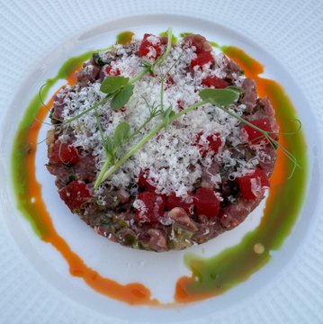 A plated dish resembling tartare is garnished with grated cheese, microgreens, and diced red toppings, drizzled with green and orange sauces in a decorative circular pattern.