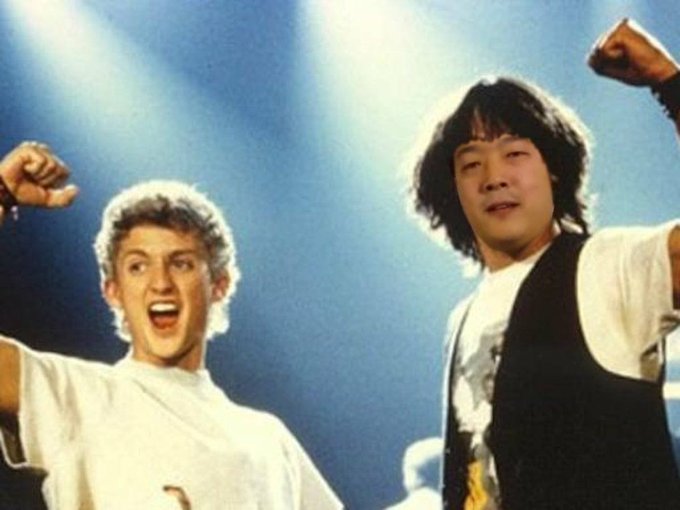 The image shows two young men, one with blonde hair wearing a white shirt and the other with dark hair wearing a black and white top, both raising their fists in an enthusiastic pose under bright stage lighting, likely from the film "Bill & Ted's Excellent Adventure" featuring characters played by Keanu Reeves and Alex Winter.