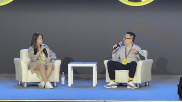 Two people seated in white armchairs on a stage during a panel discussion, with the man holding a microphone and speaking.