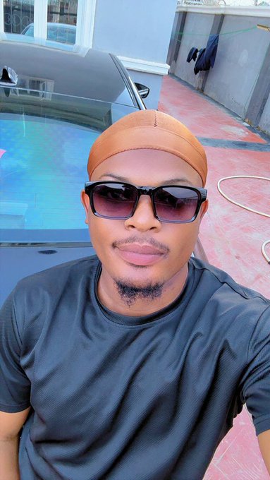 A selfie of a man wearing a brown durag, black sunglasses, and a black t-shirt, posing outdoors with a blue swimming pool and red flooring visible in the background.