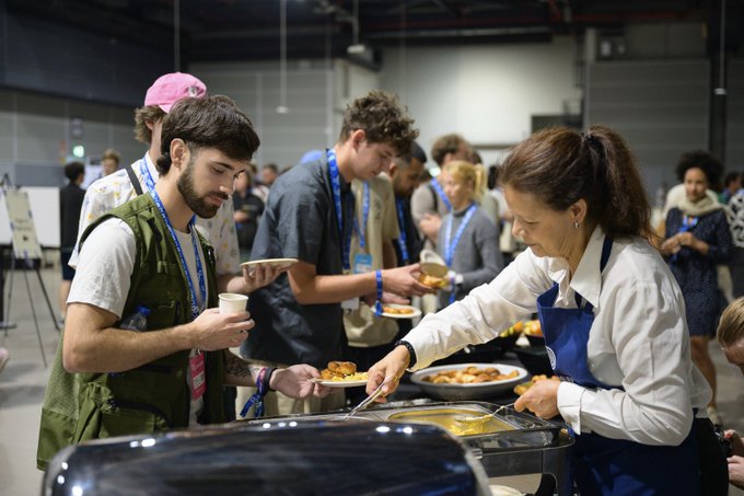 People wearing lanyards gather around a food service station where a woman in a white shirt and blue apron serves food onto plates.