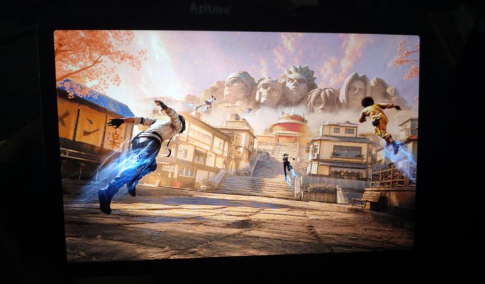 A monitor displays a fighting video game scene with characters wielding blue energy attacks in a village setting, with a large mountain carved with faces in the background.