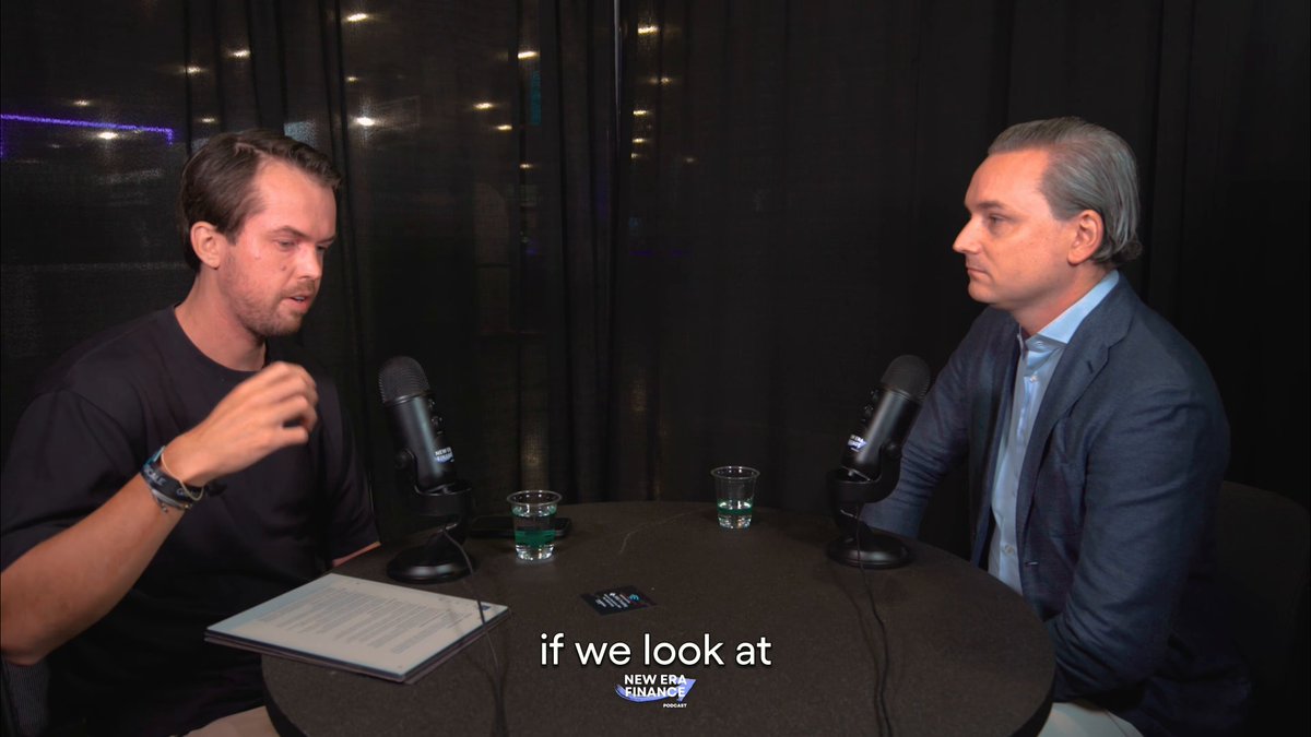 The image shows two men sitting at a table, with one man holding a microphone while the other man has a book open in front of him. The scene suggests that they are participating in an interview or discussion about tokenization and its impact on institutional capital.