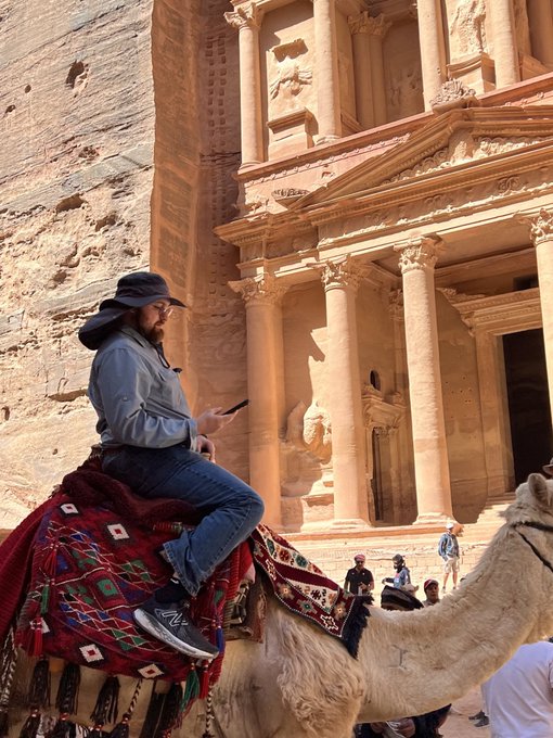 A man in a sun hat and gray shirt rides a camel past the stunning rock-cut facade of Al-Khazneh (The Treasury) in Petra, Jordan, holding his phone while tourists gather below. The image humorously captures a "blend of old and new," with the casual smartphone-wielding rider juxtaposed against the ancient Nabataean monument—perfectly embodying the viral, meme-status post.