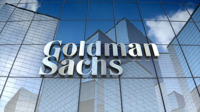The exterior of a Goldman Sachs building featuring the company's silver logo mounted on a reflective glass facade that mirrors the blue sky and clouds above.