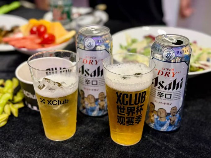 Two frosty pints of Asahi Super "Dry" beer branded with "XClub 世界杯观赛季" (World Cup viewing season) logos sit alongside their matching cans on a snack table featuring fresh fruit and salad.

The image ties to the post by showcasing the "XClub" and "OKX"-branded beer cans from what appears to be a World Cup viewing event where the author participated in a prediction contest, casting their votes for Argentina despite not winning the prize.