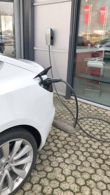 The front left side of a white Volvo is plugged into a wall-mounted EV charging station, with the charging cable draped over a paved stone surface.