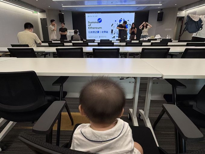 A baby with very short hair sits in an office chair facing the front of a meeting room where an "Ethereum Summer" event is being held, with a presentation screen reading "Summer of Ethereum" and "以太坊之夏 (北京站)" visible at the front, where several attendees are gathered. The image humorously illustrates the caption's suggestion that the baby may be the youngest Ethereum builder, as the child appears to be attentively watching the crypto meetup.