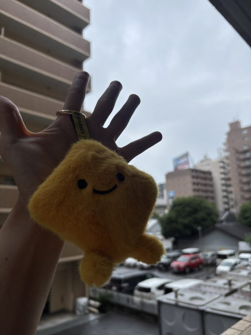 A hand holding a small fuzzy yellow plush toy with a smiley face against a backdrop of city buildings and a cloudy sky.