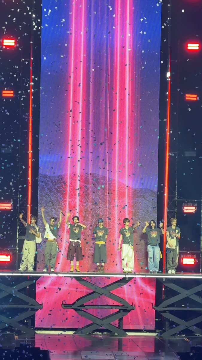 The image shows a group of people standing together, with one person holding their arms up high as if they are celebrating or cheering. The scene takes place on stage, where there is a large screen behind them that displays a vibrant pink background with red lights. The people in the photo appear to be part of an event or performance, possibly related to a music festival or concert.