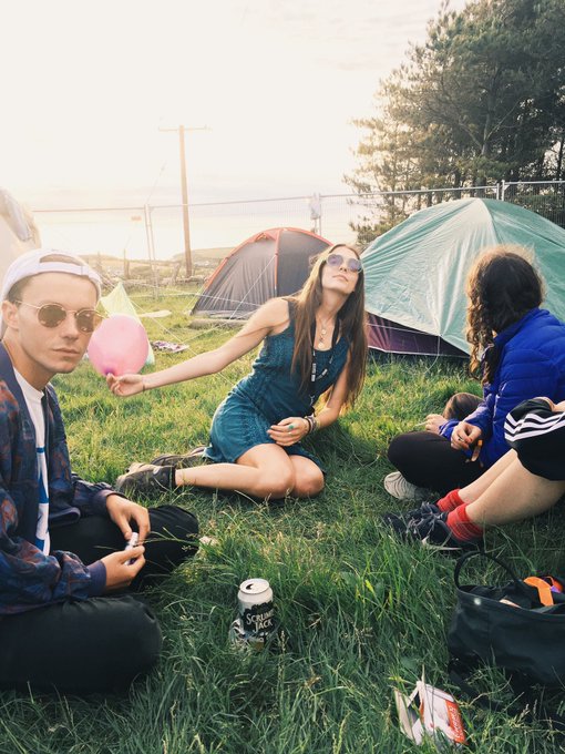 Three young people sitting in the grass at a campsite with tents in the background; one holds a pink balloon, and a can sits in the grass.
