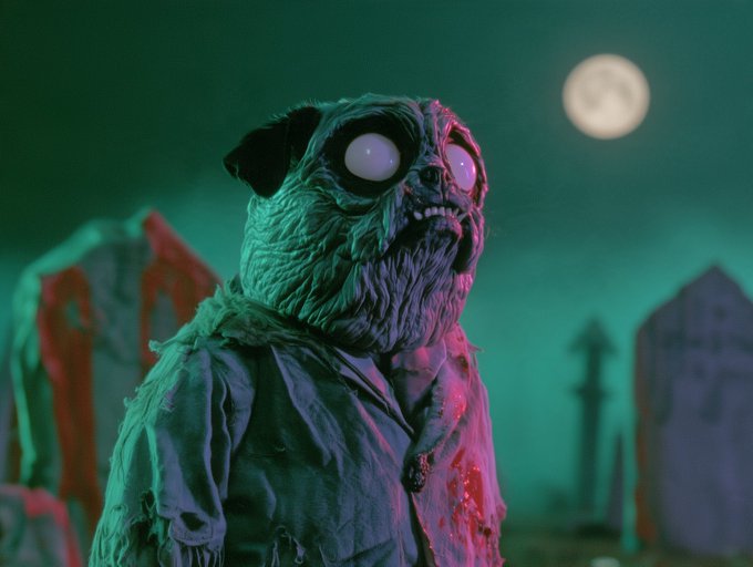 A creepy, bloodied, ghostly dog-like creature with bulging eyes and tattered clothing in a Halloween-themed scene with a full moon and tombstones in the background.