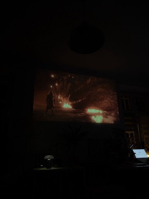 A dark room with a projector displaying a video game scene featuring a warrior character, a large dragon head, and glowing green text reading 'GANMAW YCVRR'.