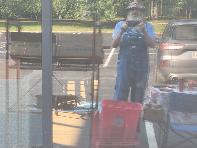An older man with a long white beard, wearing overalls and a patterned hat, stands outdoors in a parking lot taking a photo with his phone, with a metal cart, red storage bin, and parked cars visible around him.