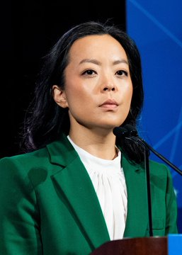 A woman with long dark hair, wearing a green blazer over a white blouse, stands at a podium with a microphone, appearing poised and attentive. The image aligns with the post about Democratic Socialist Francesca Hong's rising odds to win Wisconsin's Democratic gubernatorial nomination.