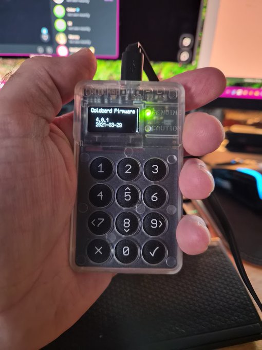 A hand holds a small keypad device labeled "TOUCHID PRO" displaying "Coldcard Firmware 5.6.1 2021-03-29," representing the cold wallet that WatchGuard will protect in an upcoming live security test.