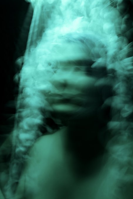 The image features a blurry photo of a person's face, with their head partially obscured by smoke or fog. The background appears to be black, which contrasts with the greenish-blue tint of the smoke. The caption above the image reads "Gm, it' s #TEZOSTUESDAY on @TeiaCommunity !" and "@TeiaWeave is LIVE! Artists on Teia have already created something beautiful." This post seems to be promoting a social media event or gathering related to weaving, as suggested by the artist's name.