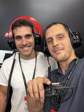 Two men wearing racing team headsets smile while holding a walkie-talkie in a garage or pit area.