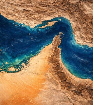 A satellite image showing the Strait of Hormuz between arid desert landmasses, with deep blue waters and a sandy coastline.