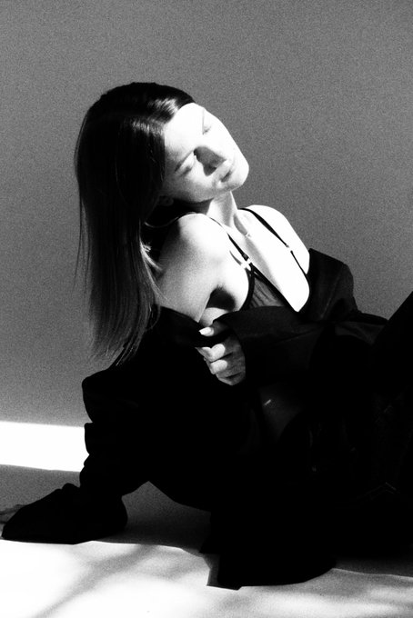 A black-and-white photograph shows a woman with long dark hair wearing a low-cut black top, leaning back with her face tilted toward the light in a dramatic pose.
