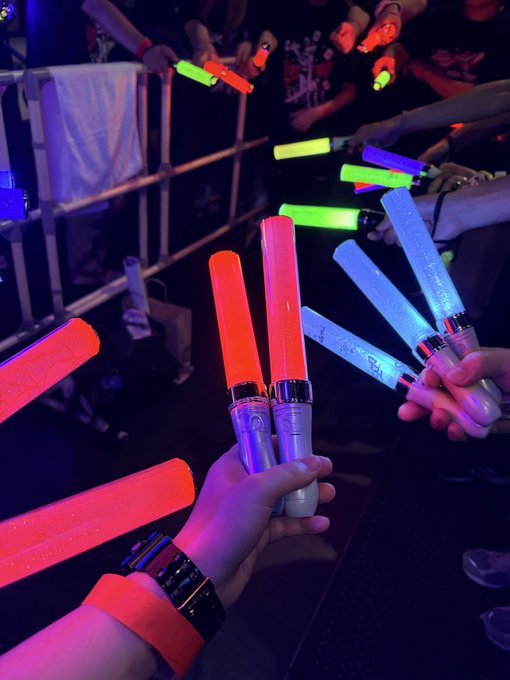 A first-person view of hands holding colorful, illuminated glow sticks, crossed with other glow sticks in a dark, concert-like environment.