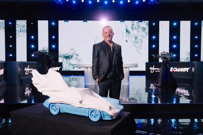 A man in a dark suit stands on a stage at a ClubWPT event, posing next to a blue car-shaped bed or display with a white cloth draped over it.