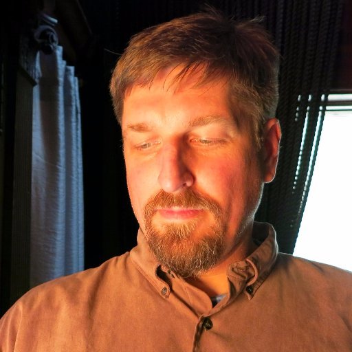 A close-up portrait of a man with short brown hair and a beard, eyes closed, illuminated by warm sunlight streaming through what appears to be a vehicle or bus window.