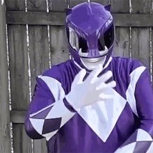 The image features a person dressed as a superhero, wearing a purple and white outfit with a helmet that has a face plate. The person's hand is raised in front of them, possibly making a gesture or posing for the photo. The background includes a wooden fence, which adds an interesting contrast to the vibrant colors of the superhero costume.