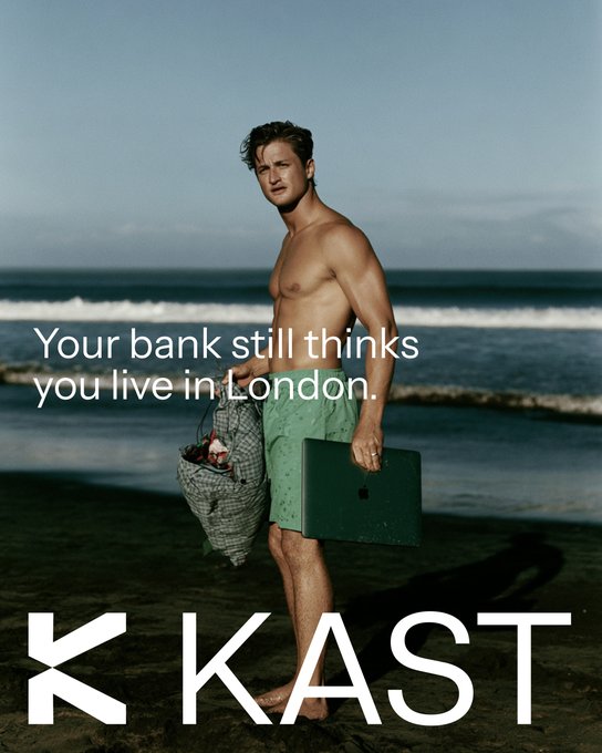 A shirtless man walks along a beach carrying a mesh bag and a laptop, with the text "Your bank still thinks you live in London" alongside the "KAST" logo. The image illustrates the post's message about needing banking services that match a nomadic lifestyle rather than a fixed address, with the man's beachside scene emphasizing life on the move while the laptop hints at remote work and digital banking.